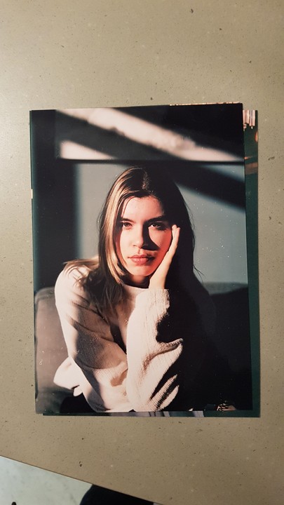 A woman with her hand on her cheek is illuminated by strong directional light creating shadows on the wall behind her.