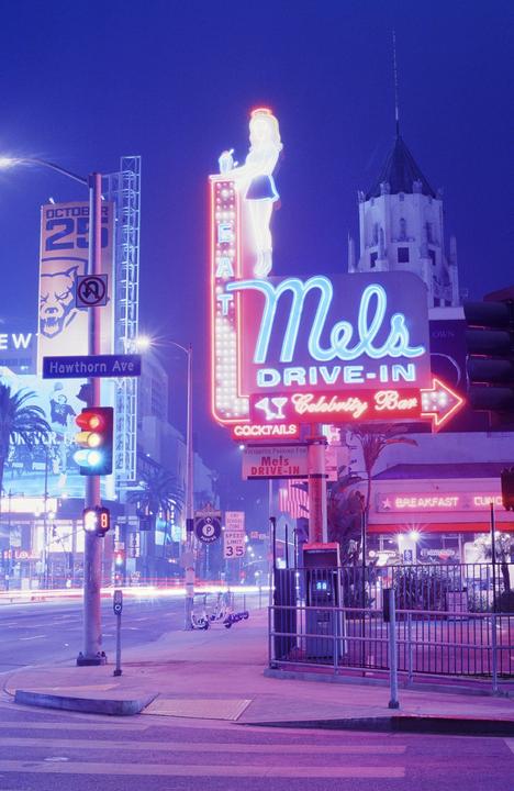 A neon sign for Mel's Drive-In with a waitress figure stands prominently on a city street at night.