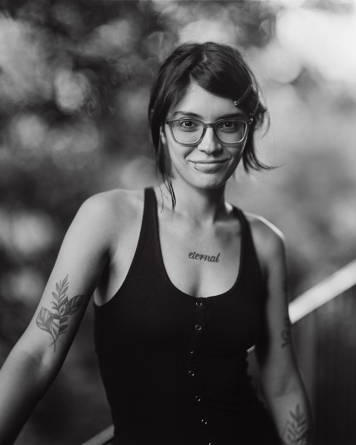 A woman with glasses and tattoos smiles at the viewer in a black and white portrait.