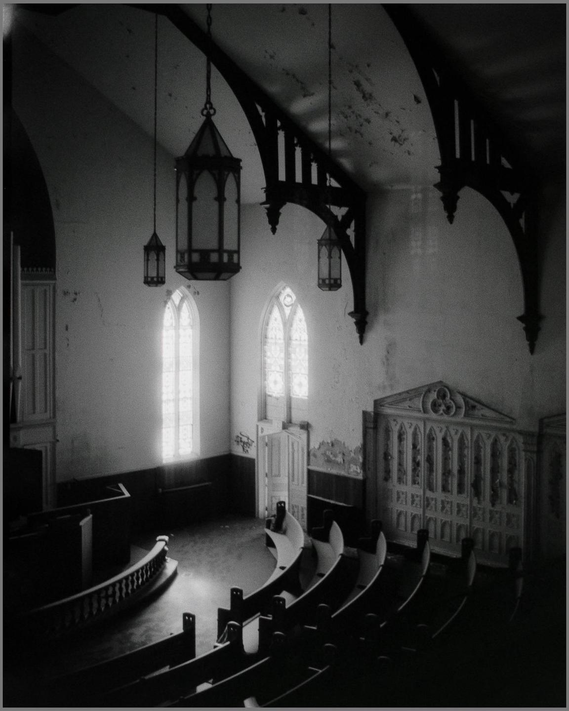 An abandoned church interior is seen from an elevated perspective, featuring arched windows, hanging lanterns, and rows of pews.