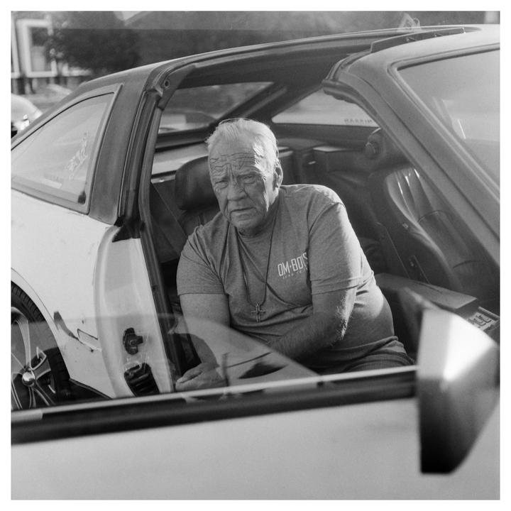 An older man with a cross necklace sits in the driver's seat of a car with the door open.