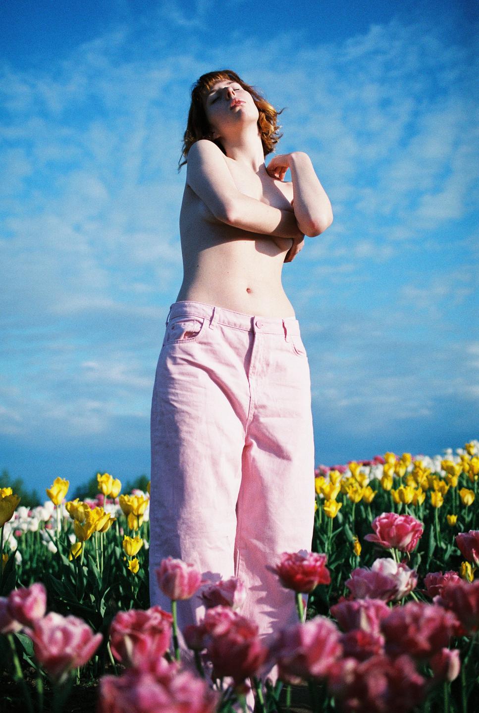 A topless woman in pink pants stands in a field of colorful tulips under a blue sky.