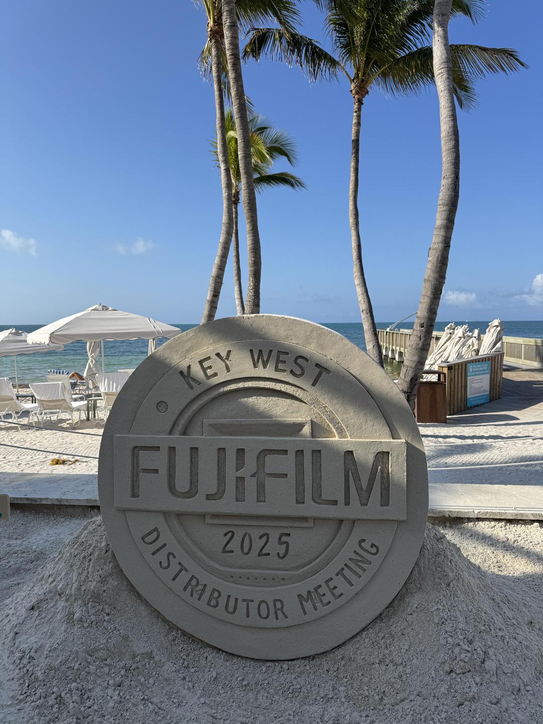 A sand sculpture with text is on a beach with palm trees and the ocean in the background.