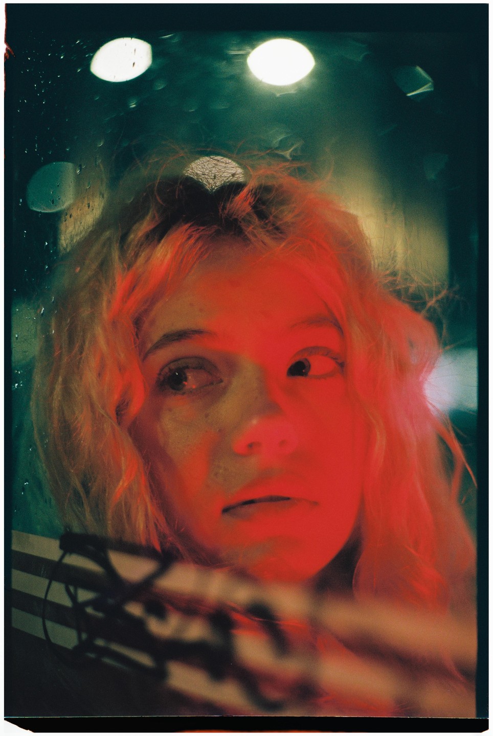 A woman with red hair and red light on her face looks out through a wet window at night.