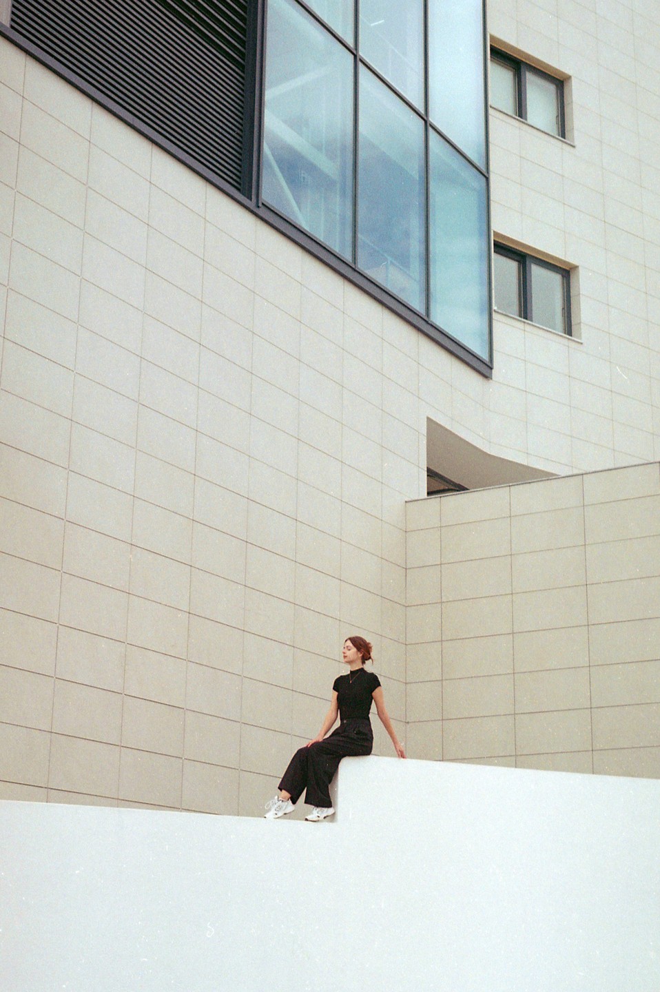 A woman in black clothing sits on a white ledge in front of a modern building with large glass windows.