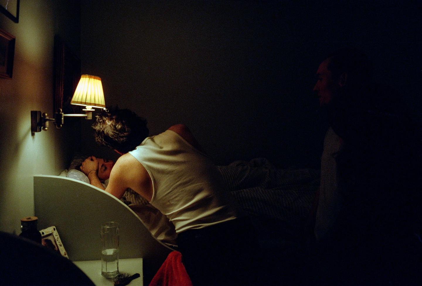 A man leans over a woman in bed, illuminated by a bedside lamp, while another man sits in shadow.
