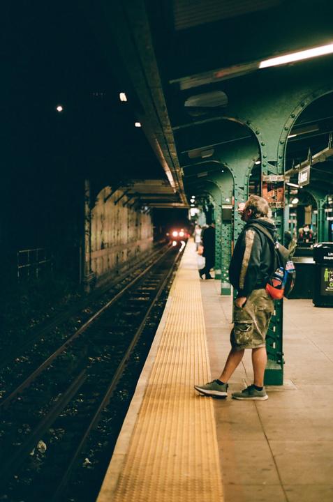 A man stands on a subway platform at night, looking down the tracks as a train approaches.
