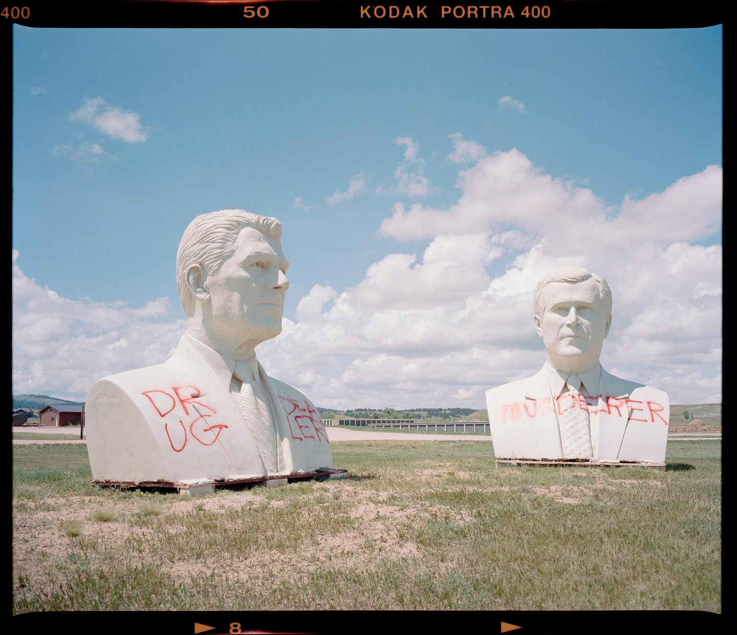 Two large white busts of men with red graffiti on them stand in a grassy field under a blue sky with white clouds.