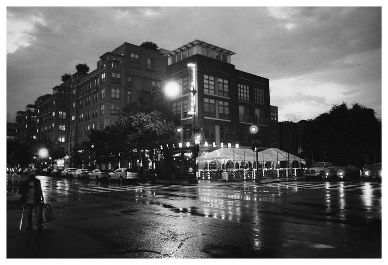 A person walks on a wet street at night with buildings and cars visible.