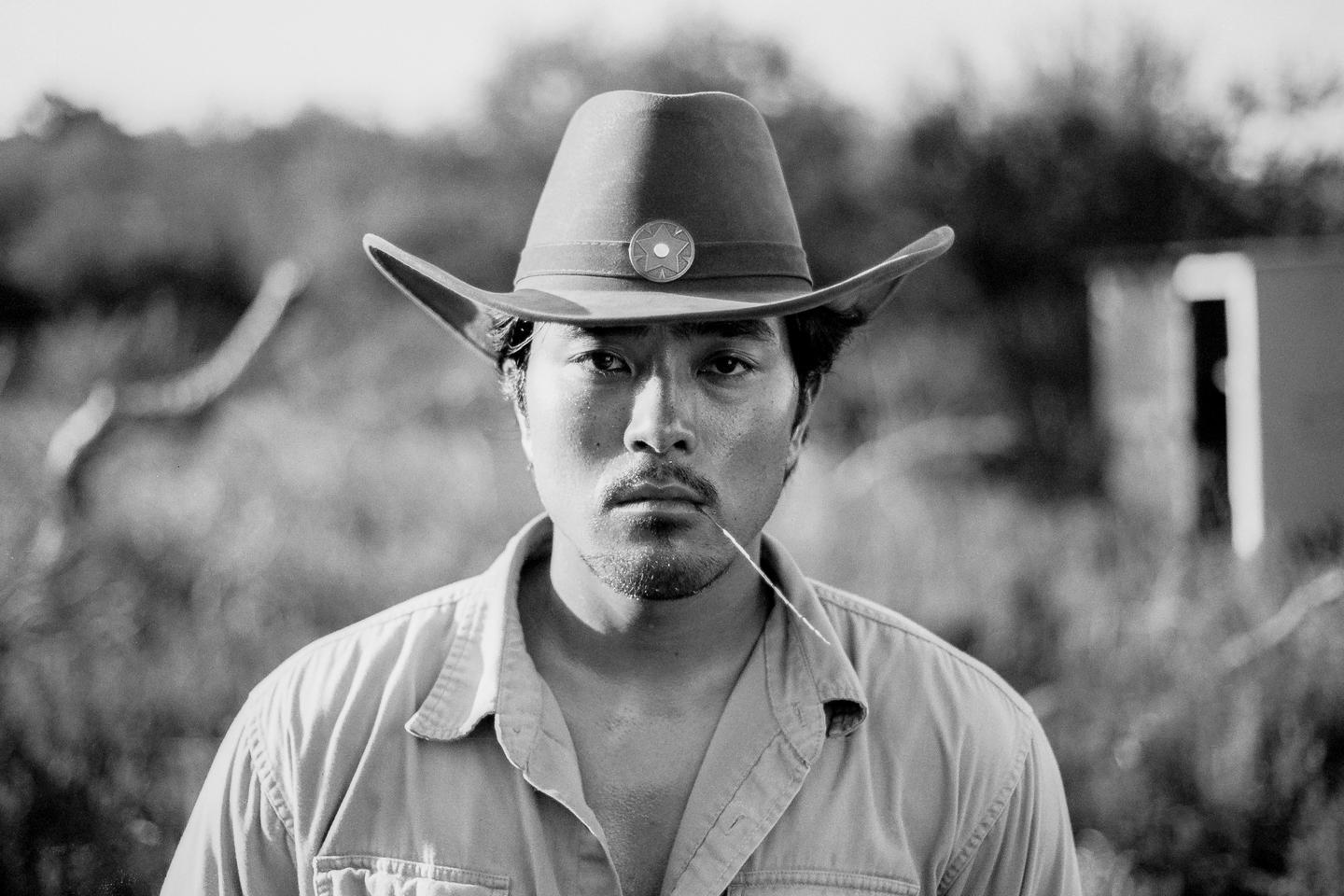 A man wearing a cowboy hat and a shirt with an open collar has a piece of straw in his mouth.