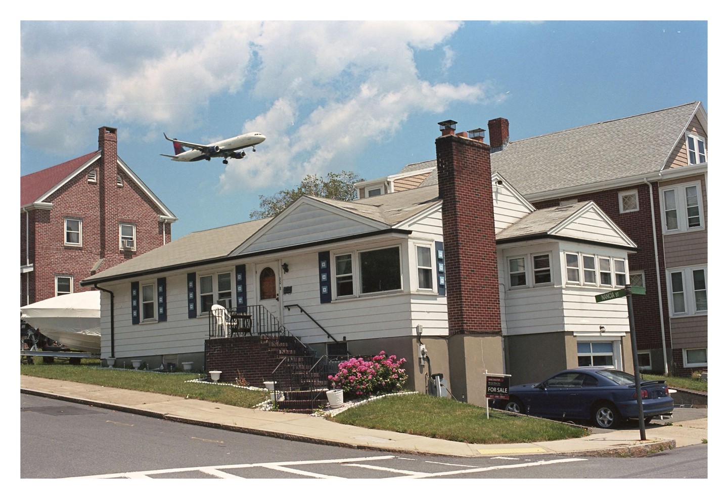 An airplane flies over a residential street with houses, a boat, and a car.
