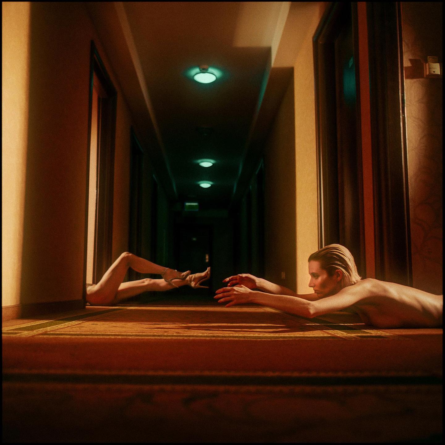 A woman lies on the floor of a hotel hallway, reaching out to a pair of legs emerging from a doorway.