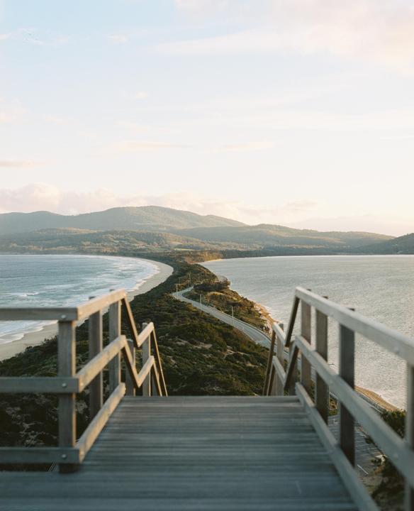 A wooden boardwalk and stairs lead down to a narrow strip of land with a road and beaches on either side, with mountains in the distance.