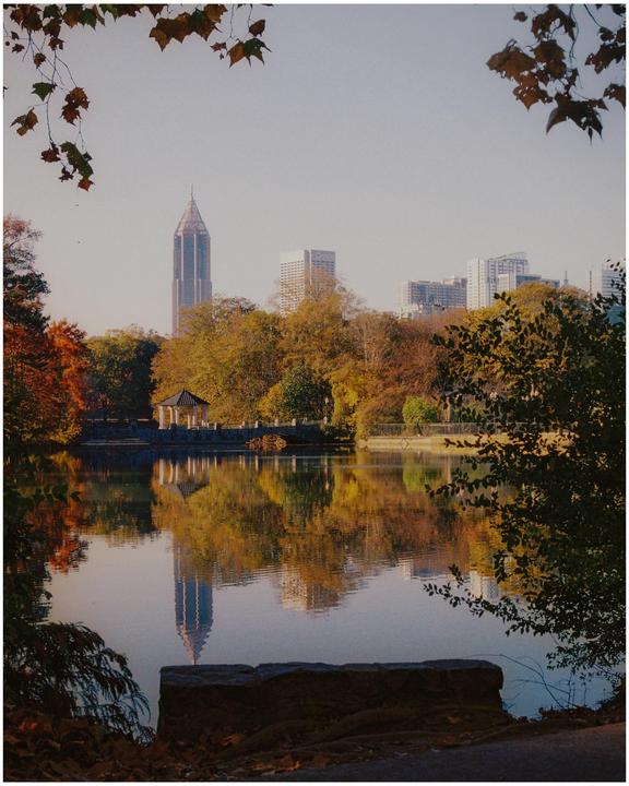 A city skyline with a prominent skyscraper is reflected in a calm lake surrounded by autumn trees and a gazebo.