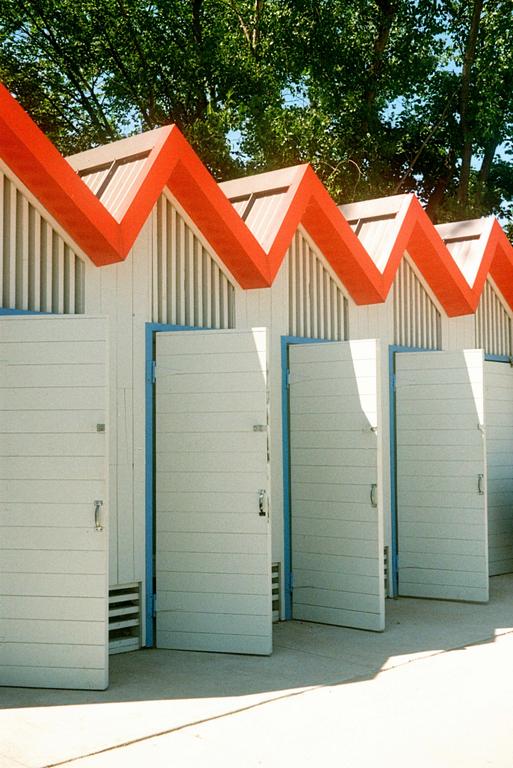 A row of white changing rooms with orange zigzag roofs stands under green trees.