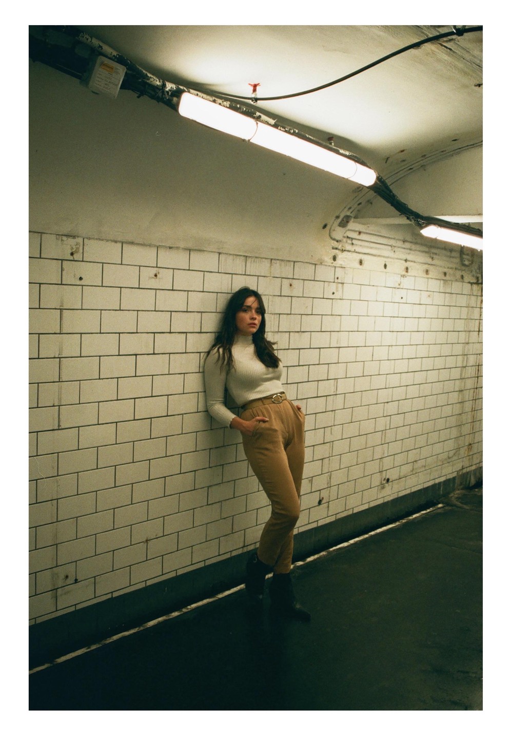 A woman with dark hair leans against a white tiled wall in a subway tunnel.