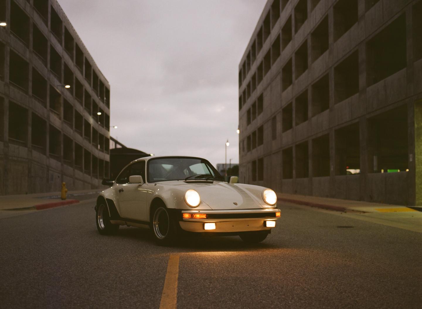 A white Porsche with its headlights on is parked on a street between two large concrete buildings.