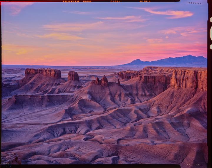 A vast desert landscape with mesas and mountains is illuminated by the colorful light of sunset.