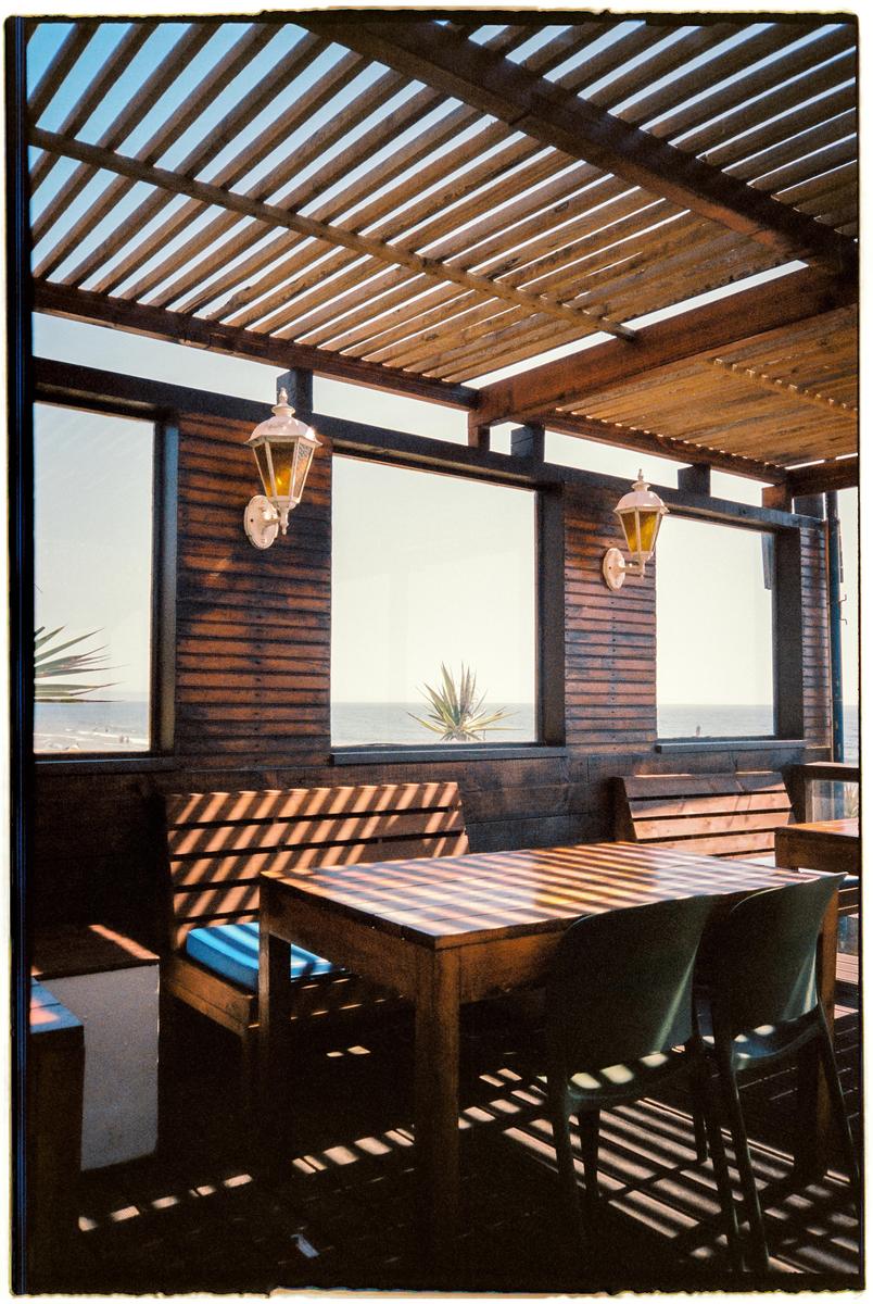 A wooden patio with a slatted roof and furniture is illuminated by strong shadows, with the ocean visible through windows in the background.