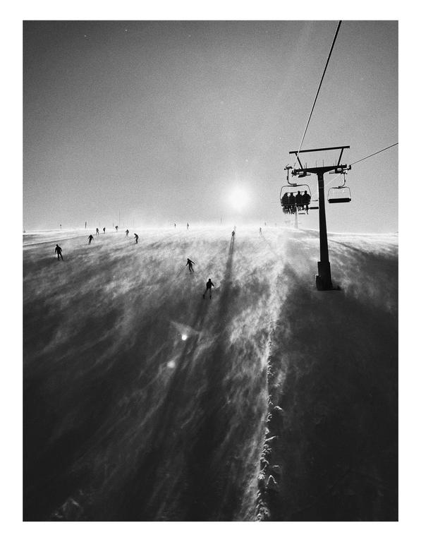 A ski lift carries people up a snowy mountain while skiers descend the slope below a bright sun.