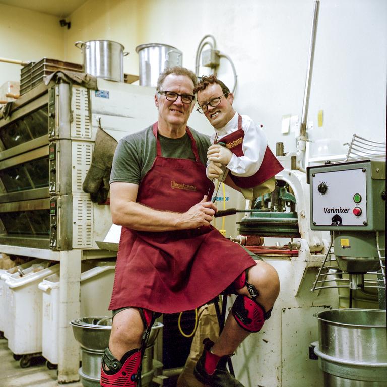 A man in an apron and knee braces sits next to a puppet in a commercial kitchen.