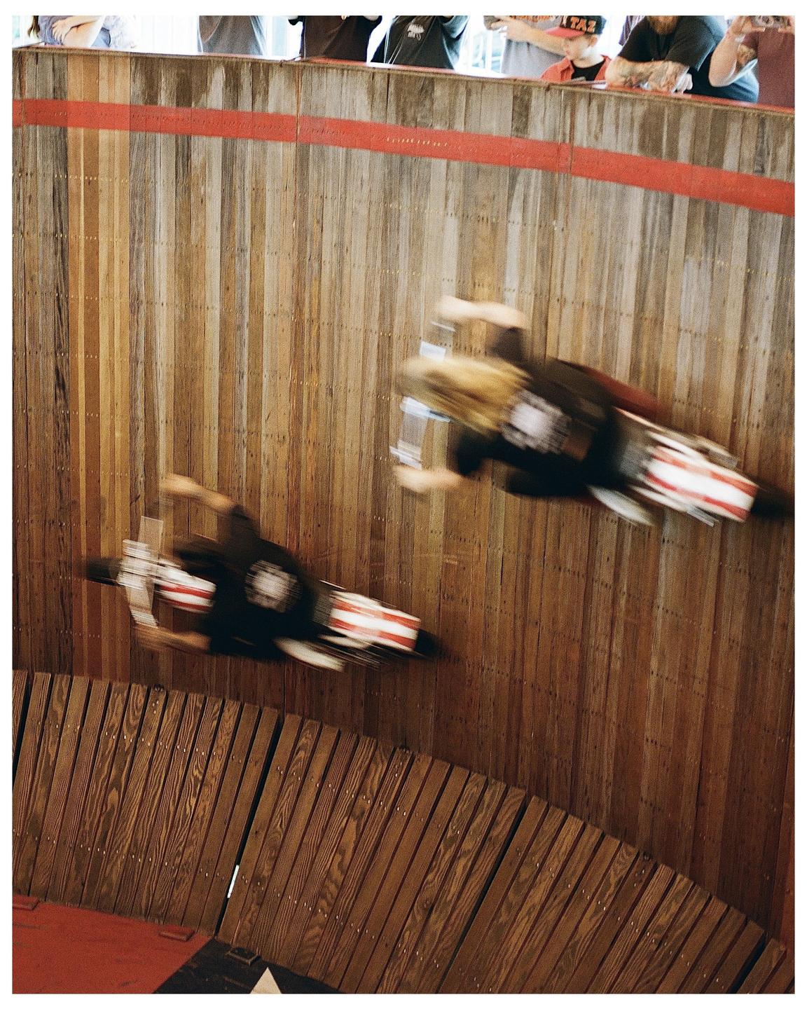 Two people ride motorcycles on a wooden wall while spectators watch from above.