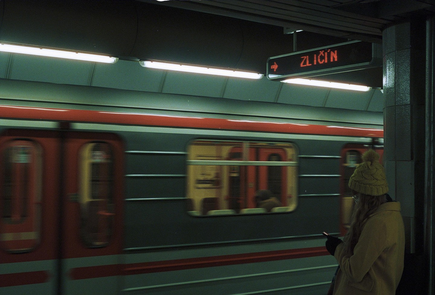 A woman in a yellow coat and hat stands on a subway platform looking at her phone as a train passes by.