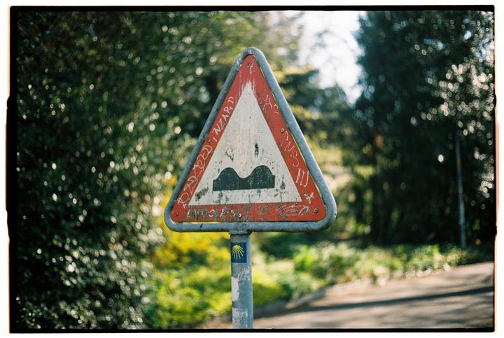 A triangular road sign indicating a bumpy road is covered in graffiti and mounted on a pole with a scallop shell sticker, set against a blurred background of green foliage.