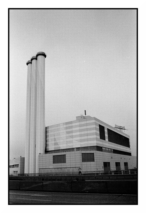 Three tall, cylindrical smokestacks stand next to a large, modern building with a patterned facade, viewed from across a road.