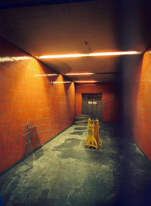 A long, narrow, orange-tiled hallway leads to an elevator, with yellow barriers and a metal grate on the floor.