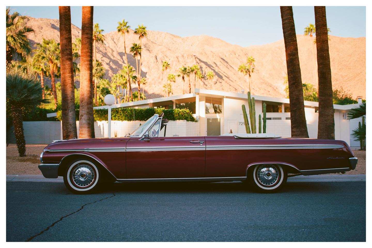 A red convertible car is parked on a street in front of a white building with palm trees and mountains in the background.