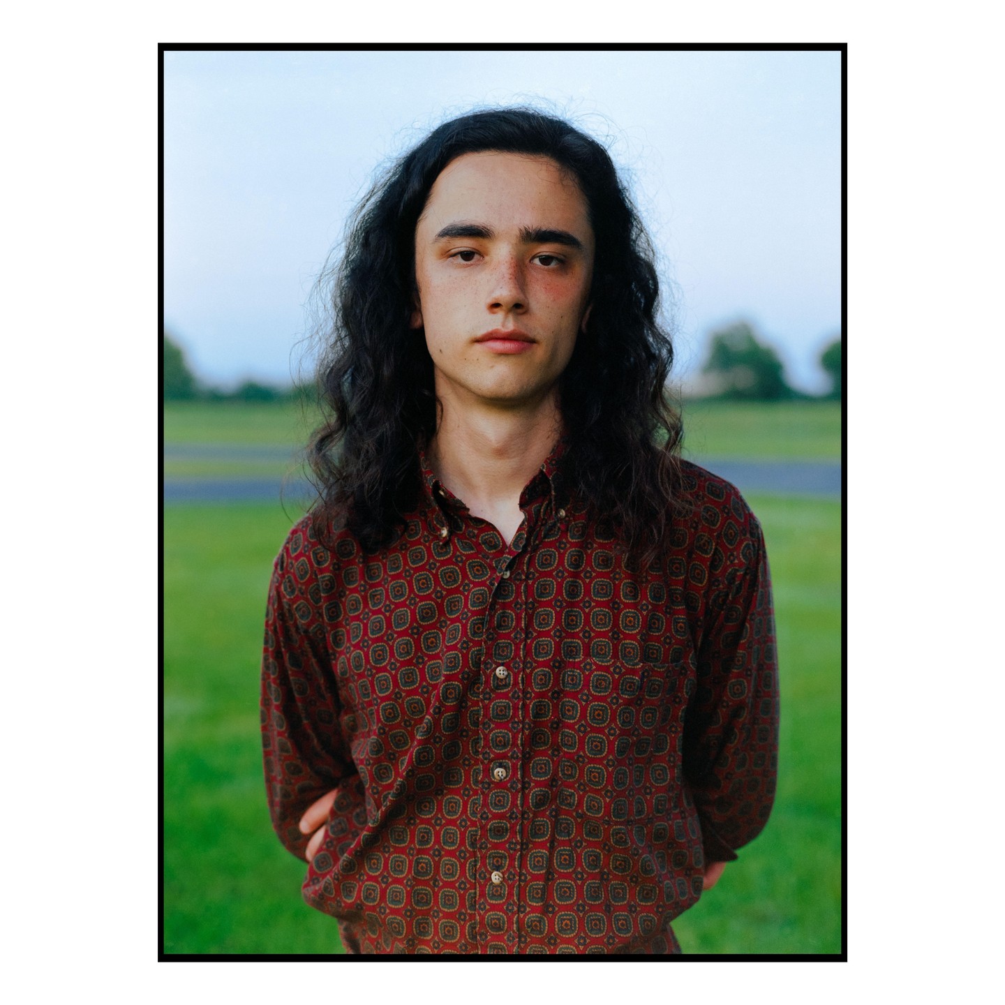 A man with long, dark, curly hair and a patterned shirt stands in a grassy field.