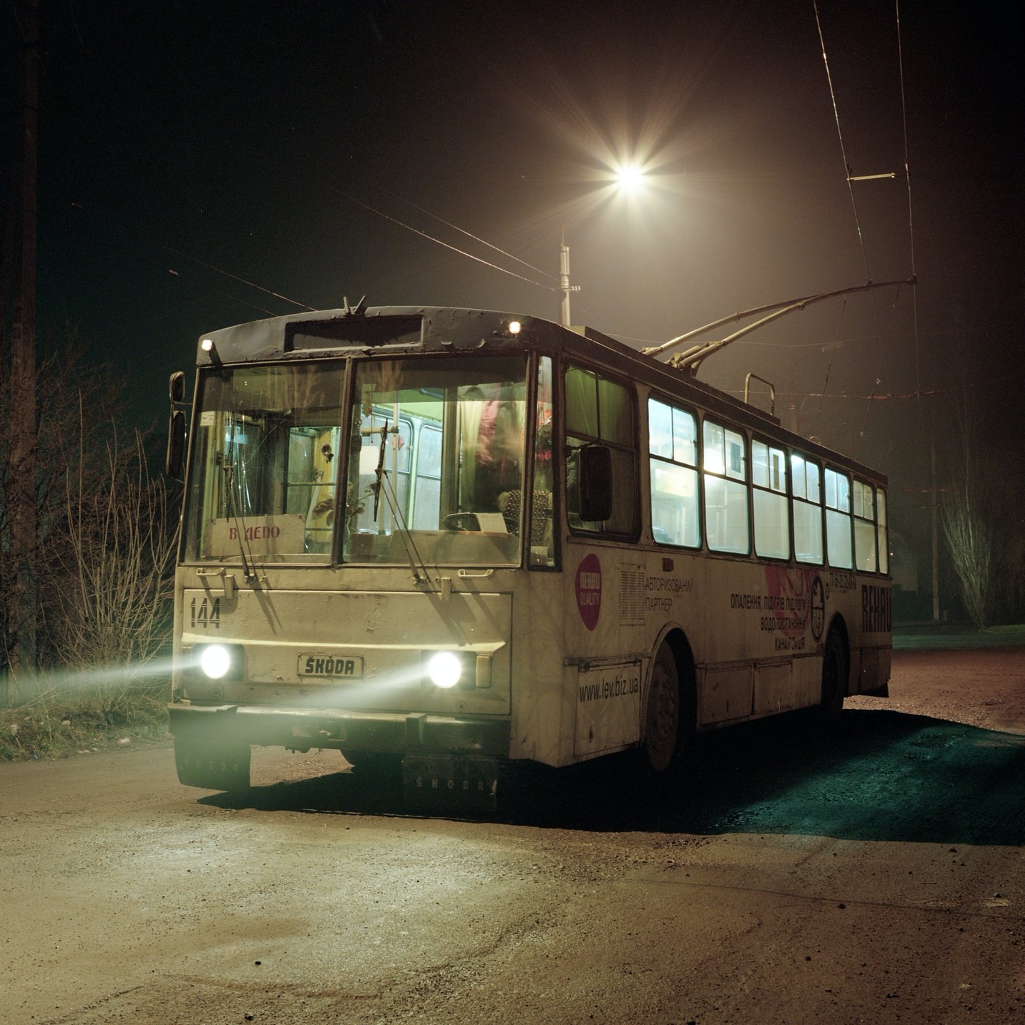 A trolleybus with its headlights on is parked on a street at night.