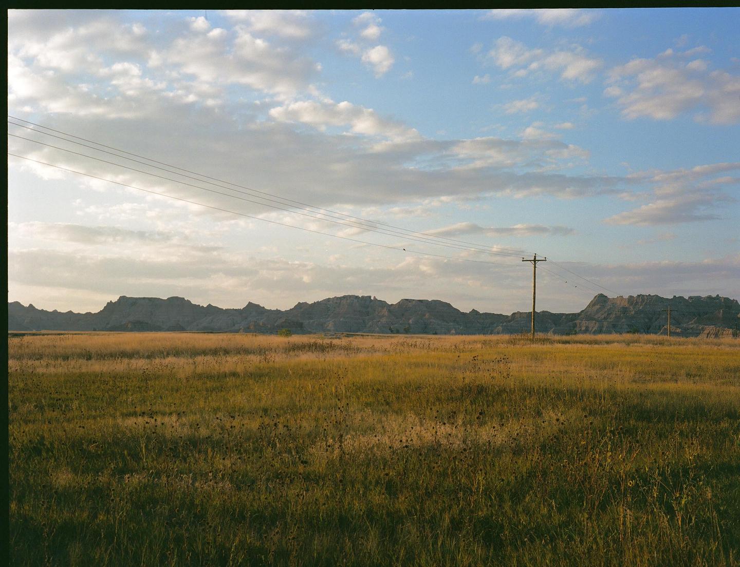 A vast field of tall grass stretches towards a distant mountain range under a cloudy sky, with power lines and a utility pole visible.