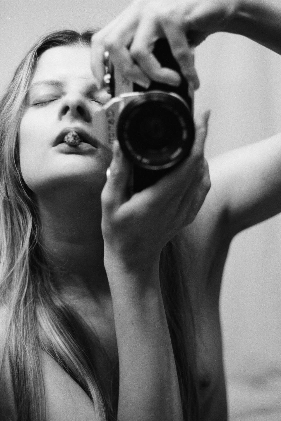 A woman with closed eyes holds a camera to take a self-portrait, with a small object on her lip.