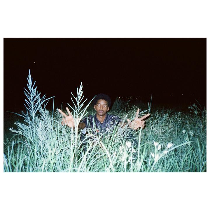 A young man crouches in tall grass at night, holding up both hands with fingers spread.
