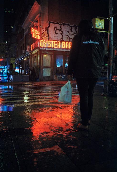A person walks across a wet street at night, past an oyster bar with neon signs.