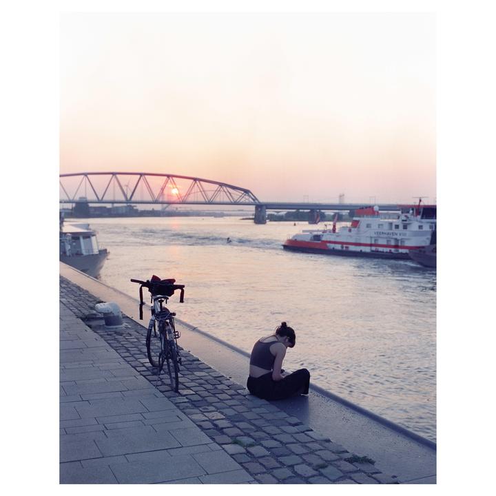 A woman sits on a cobblestone riverbank next to a bicycle, with a bridge and boats in the background at sunset.