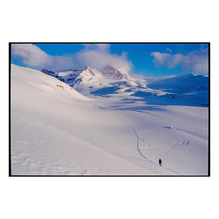 A person skis across a vast snow-covered landscape with mountains in the background under a blue sky.