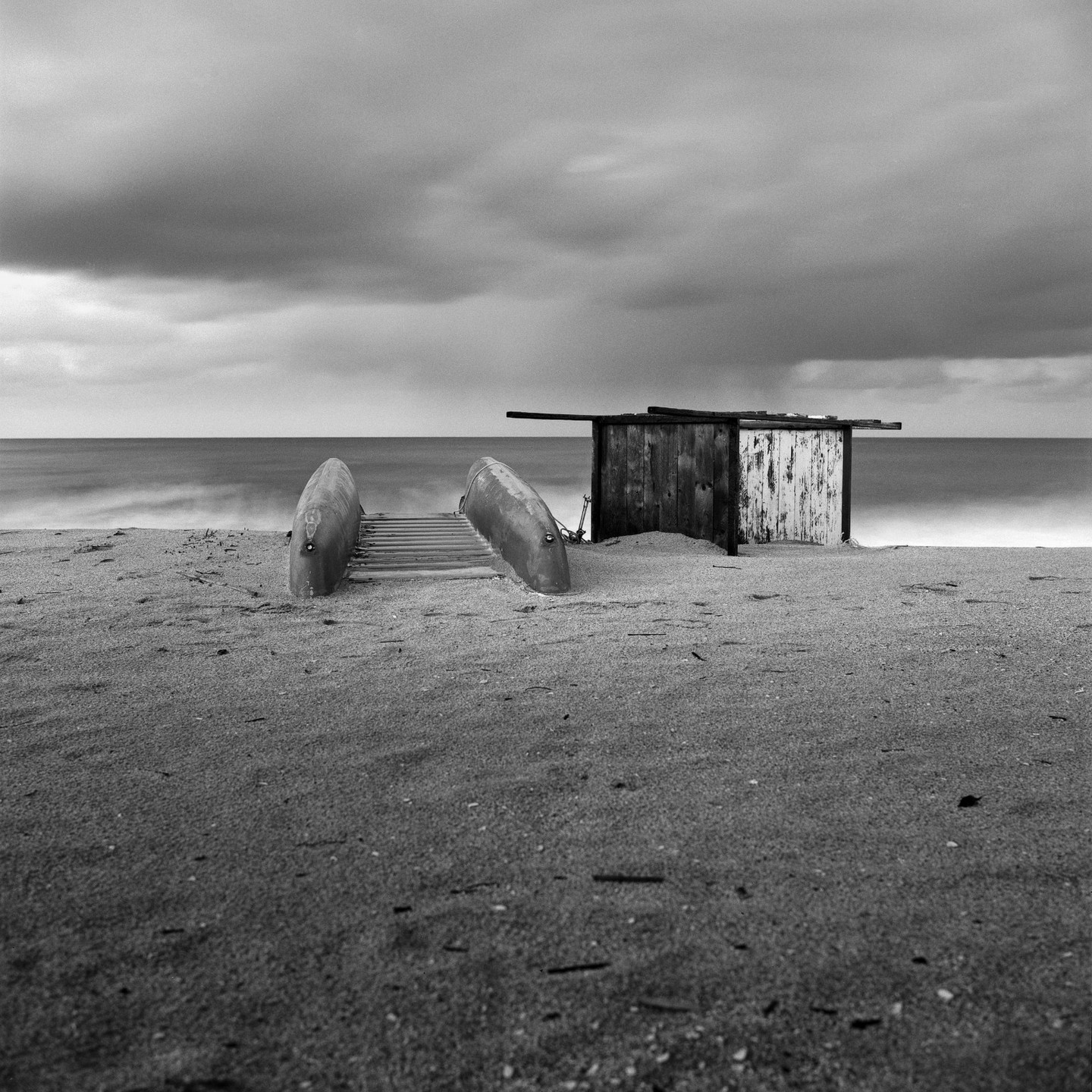 A wooden shack and two overturned boats sit on a sandy beach under a cloudy sky.