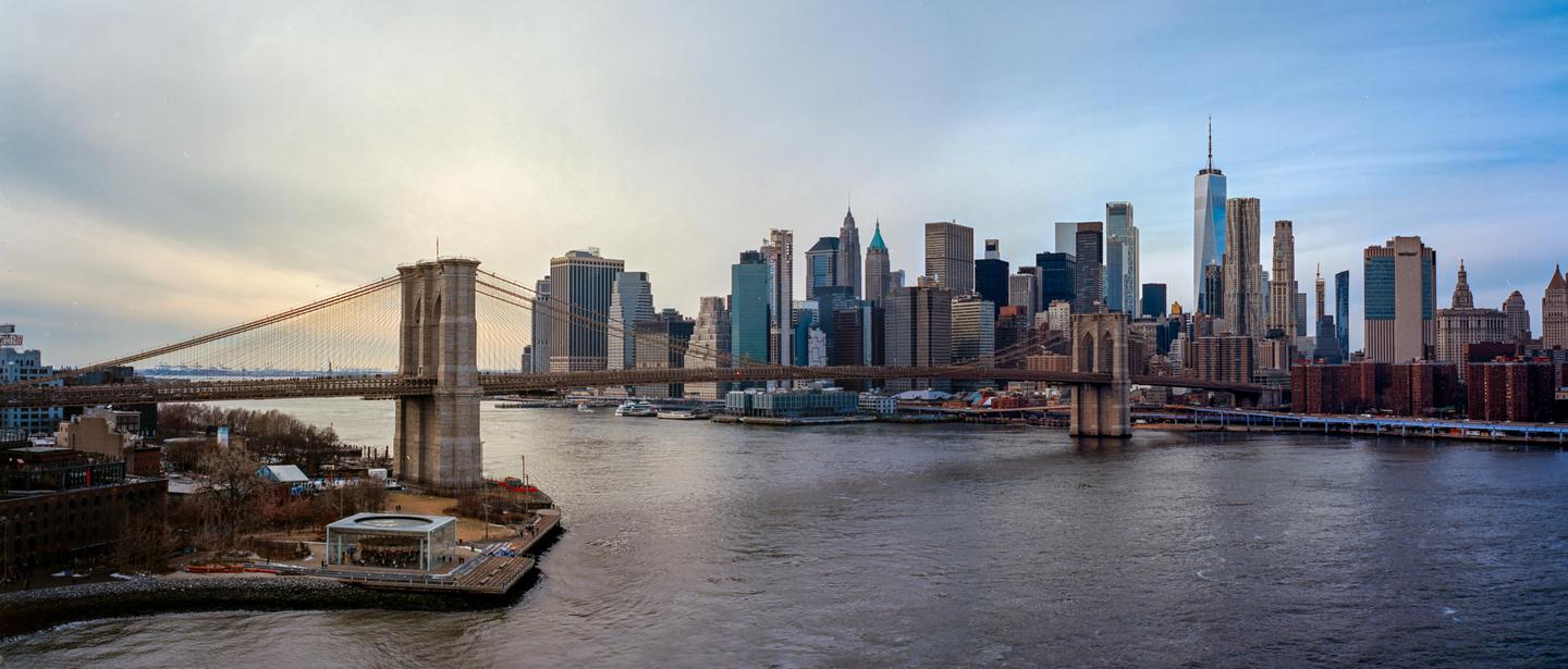 A panoramic view shows the Brooklyn Bridge spanning a river with the New York City skyline in the background.