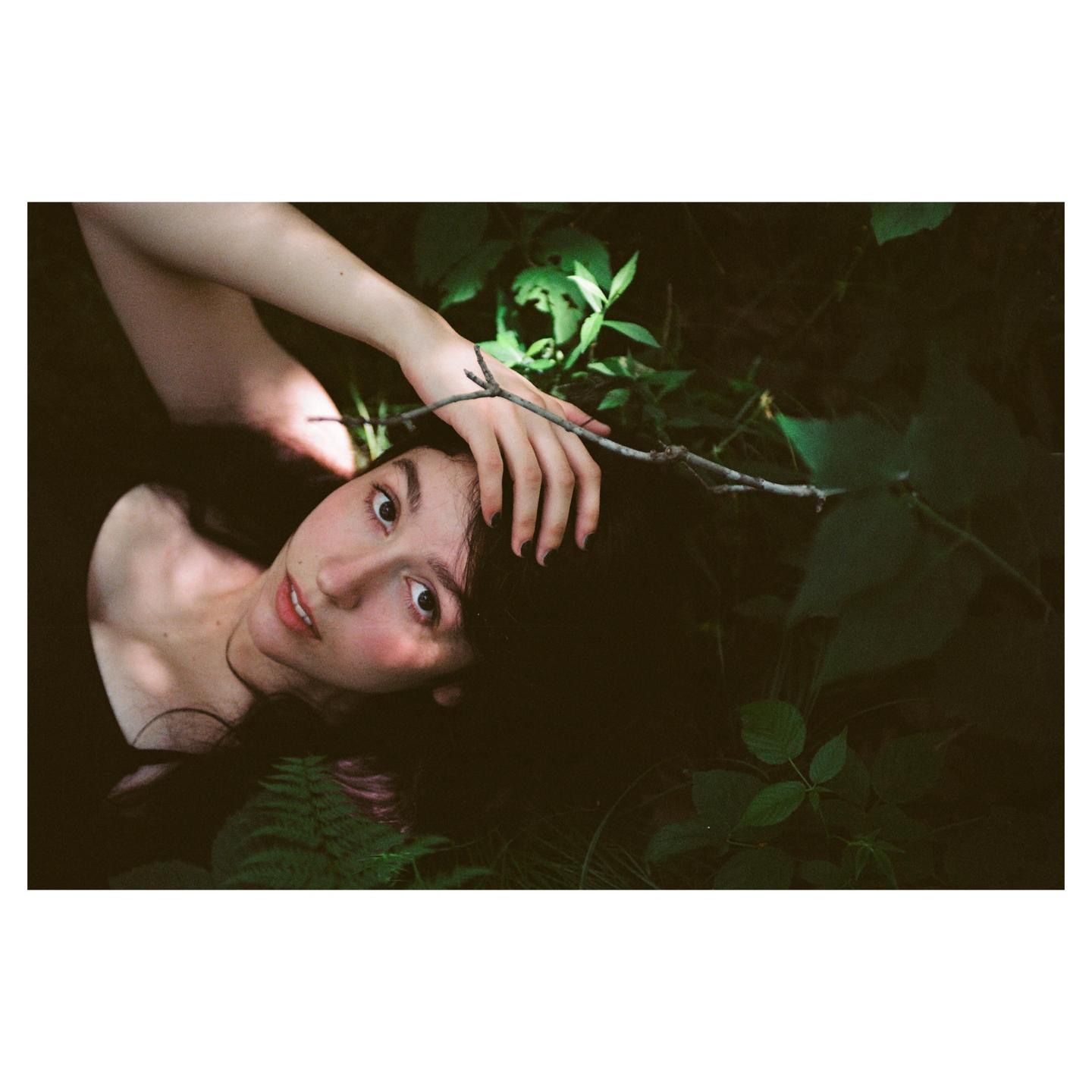 A woman with dark hair lies in a forest with her hand resting on a branch.