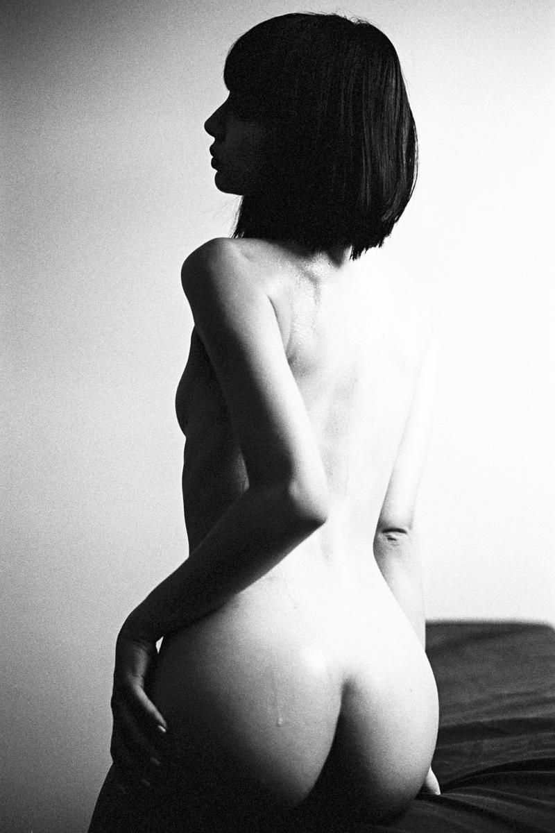 A woman with short hair is shown from behind, looking to the left, with a single drop of liquid visible on her left buttock.