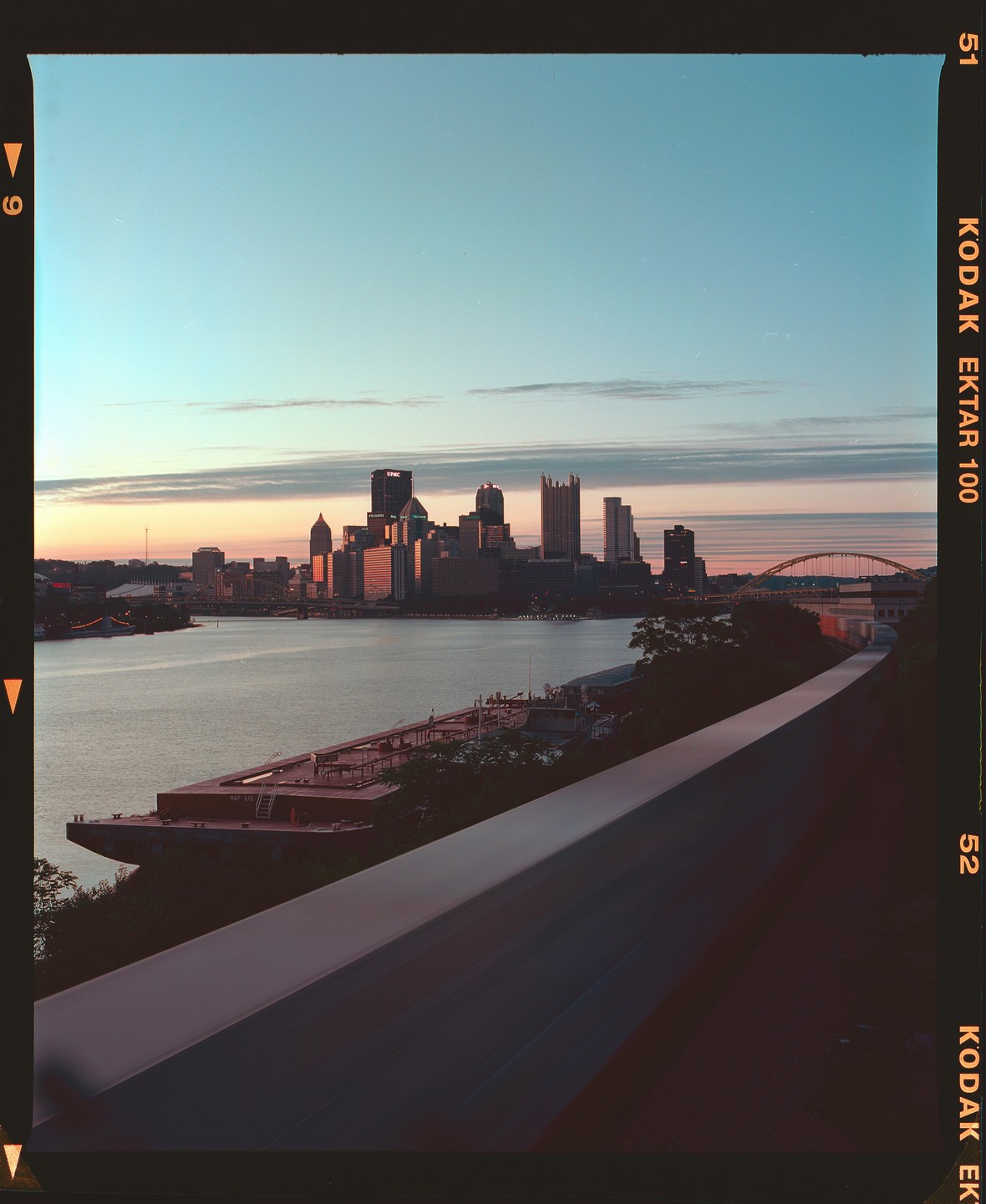 A cityscape of Pittsburgh is visible across a river with a barge in the foreground during sunset.