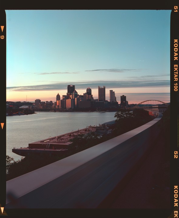 A cityscape of Pittsburgh is visible across a river with a barge in the foreground during sunset.