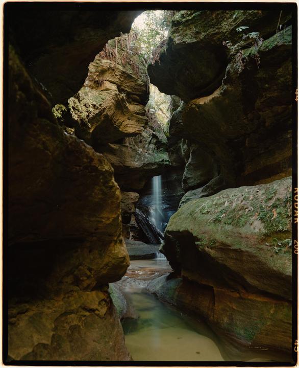 A narrow grotto with rock formations and a small waterfall flowing into a shallow stream.
