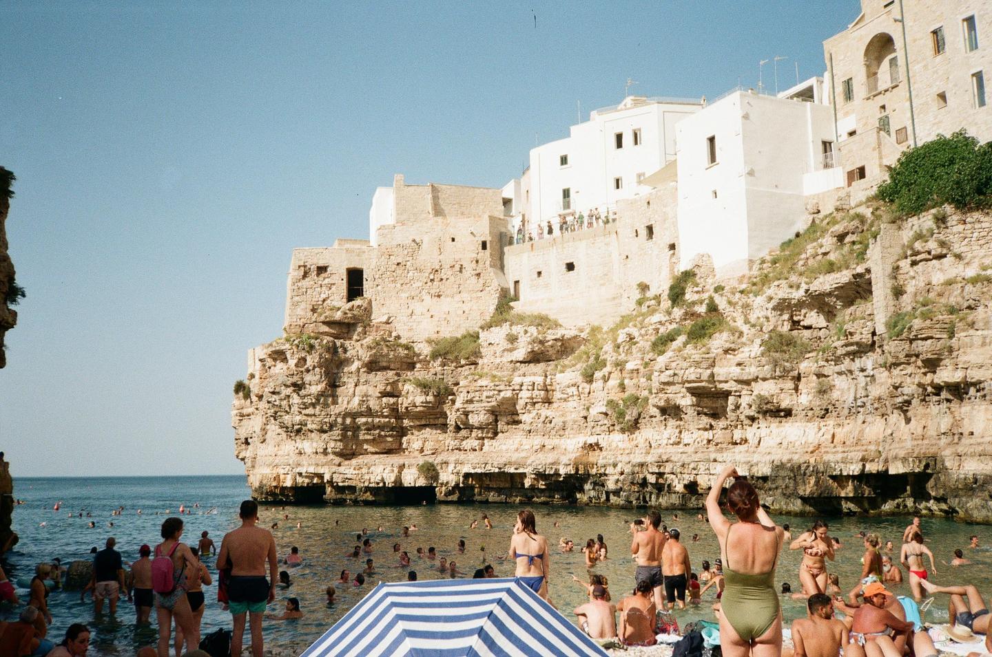 A crowd of people swim and relax on a beach next to a cliffside town.