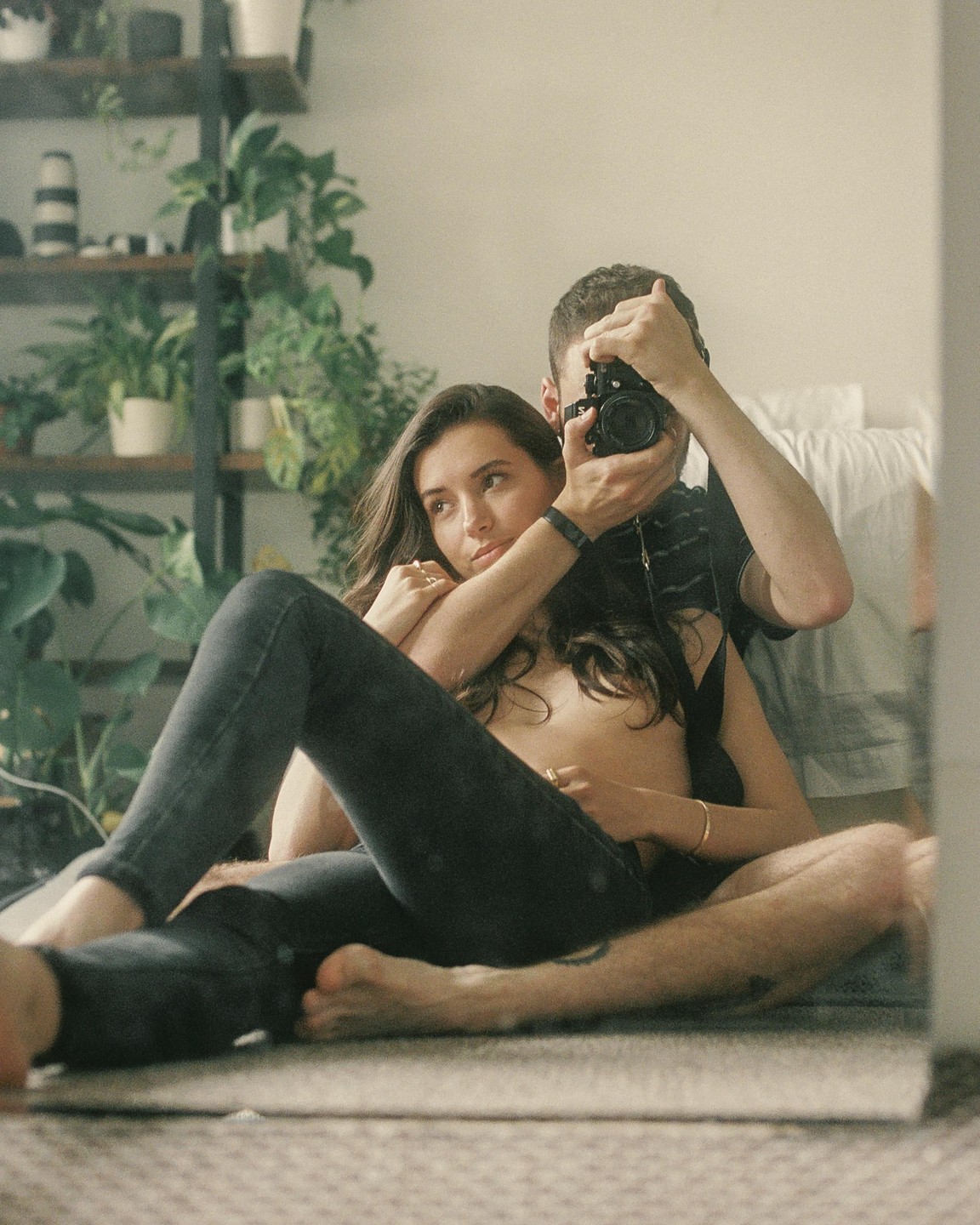 A man photographs a woman in a mirror, with plants in the background.