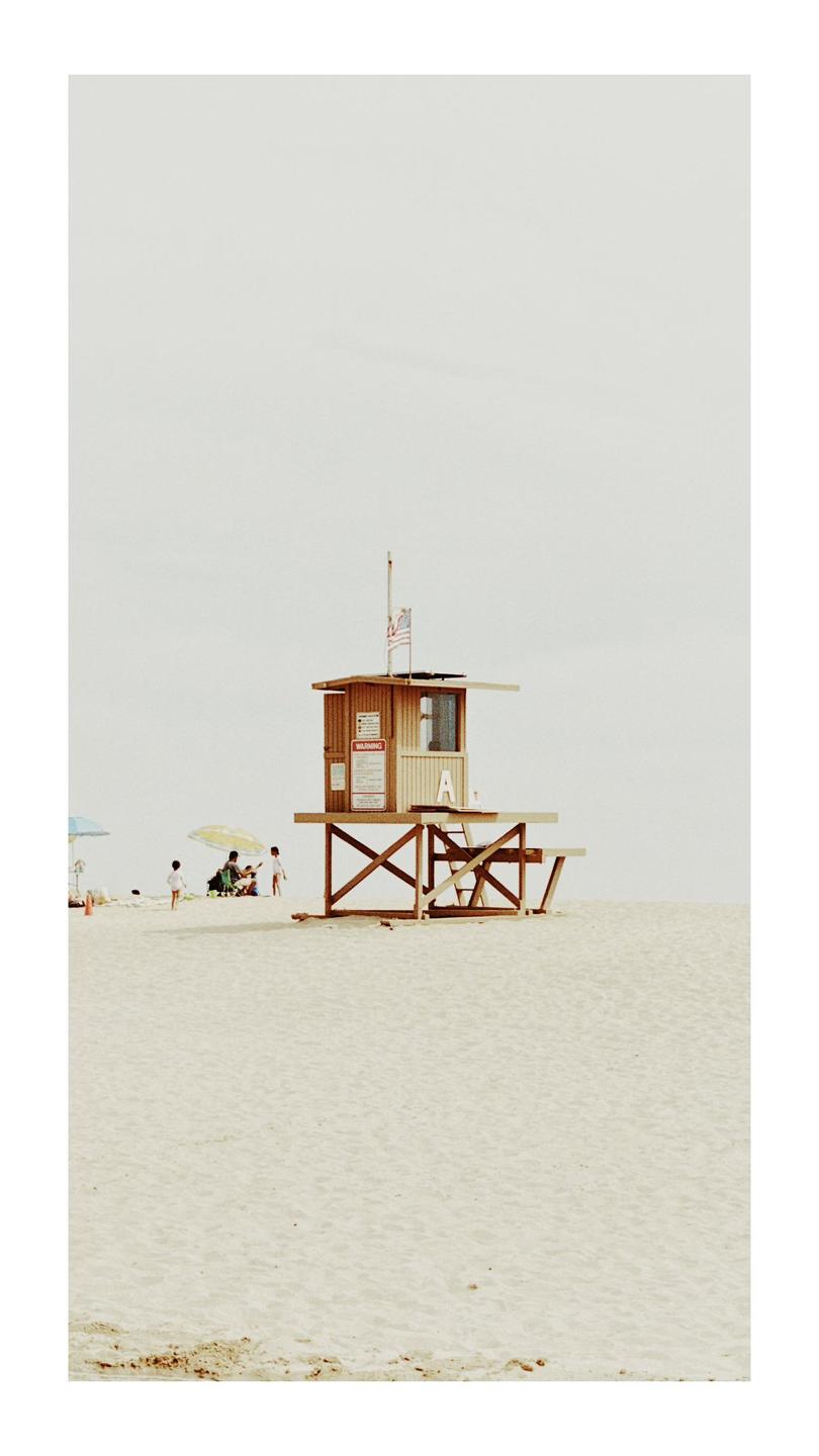 A wooden lifeguard station stands on a sandy beach with people and umbrellas in the distance.