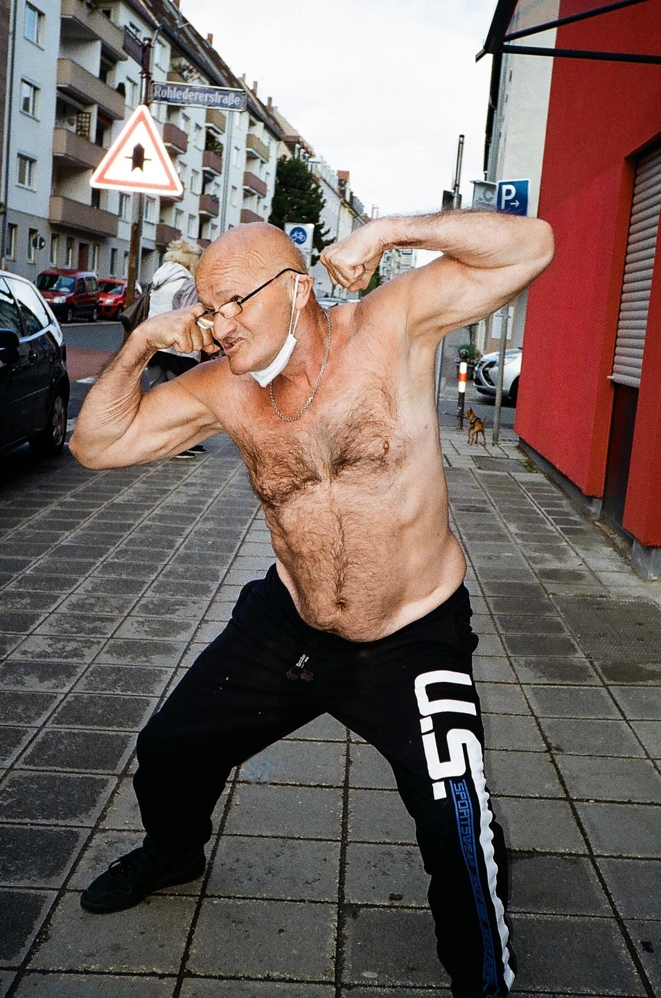 A shirtless man flexes his muscles on a city street.